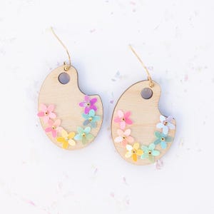 May include: A pair of wooden earrings shaped like artist's palettes, decorated with colorful flowers. The earrings are made of light wood and have a gold-colored wire.