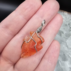 May include: A carnelian arrowhead pendant wrapped in silver-colored wire. The orange-red stone has a natural, textured appearance. The wire is looped at the top for a necklace chain. The pendant is held in a hand.