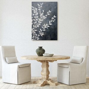 May include: A white dining room with a round wooden table and two white chairs. A black and white abstract painting hangs on the wall above the table. A wooden cabinet with glass shelves is in the background.
