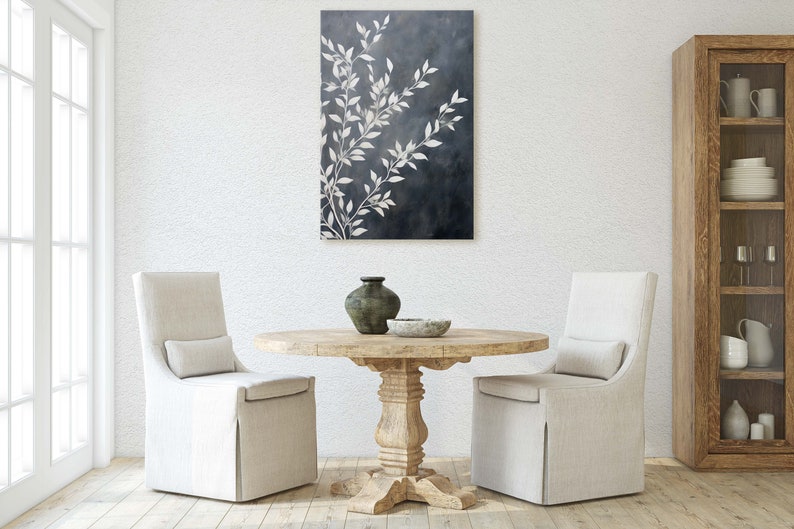 May include: A white dining room with a round wooden table and two white chairs. A black and white abstract painting hangs on the wall above the table. A wooden cabinet with glass shelves is in the background.