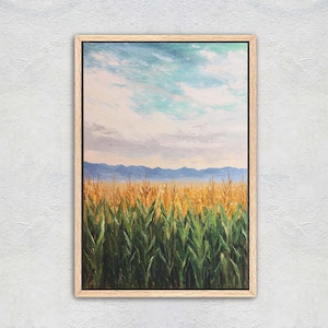May include: A framed landscape painting depicts a field of corn under a cloudy sky. The painting features a vibrant color palette with green, yellow, blue, and white hues. The artwork is encased in a light brown wooden frame.