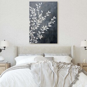 May include: A white bed with a white and gray blanket and two white lamps on the nightstands. A framed black and white photo is on the left nightstand. A painting of white branches on a dark blue background hangs above the bed.