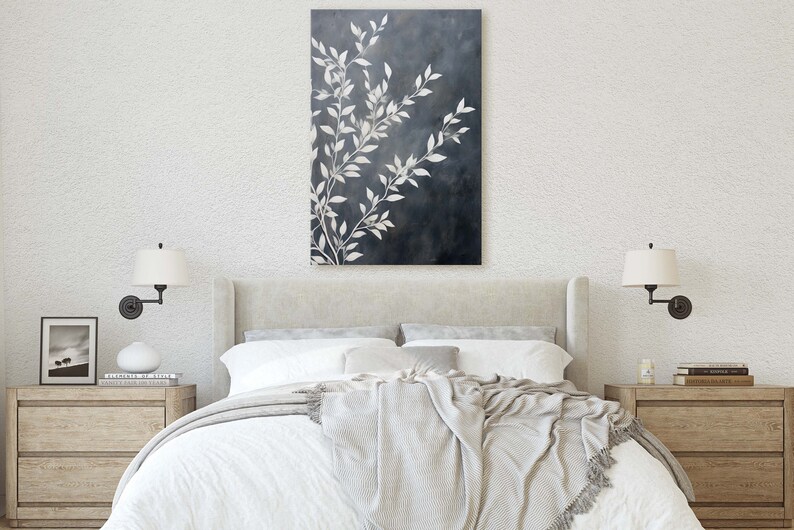 May include: A white bed with a white and gray blanket and two white lamps on the nightstands. A framed black and white photo is on the left nightstand. A painting of white branches on a dark blue background hangs above the bed.