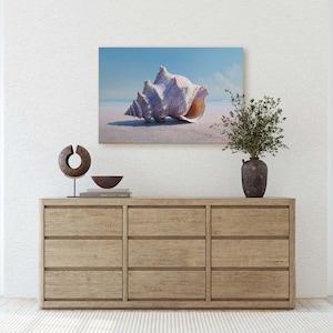 May include: A large, light-colored seashell painting hangs above a wooden dresser. The painting depicts a seashell on a sandy beach with a blue sky. A decorative sculpture and a small bowl sit on the dresser, along with a vase of greenery.