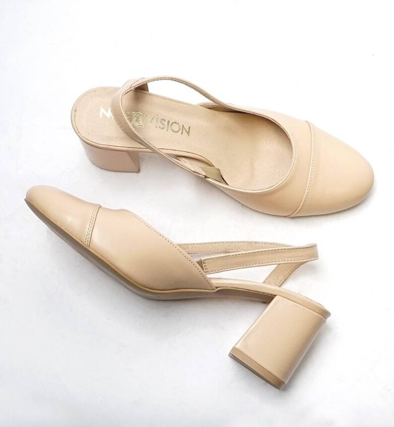May include: A pair of beige heeled slingback shoes with a block heel. The shoes have a strap that goes around the ankle and a buckle closure. The brand name "My Vision" is printed on the insole.