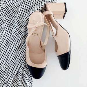 May include: A pair of beige heeled shoes with black toe caps and ankle straps. The shoes are on a white surface with a black and white polka dot fabric.