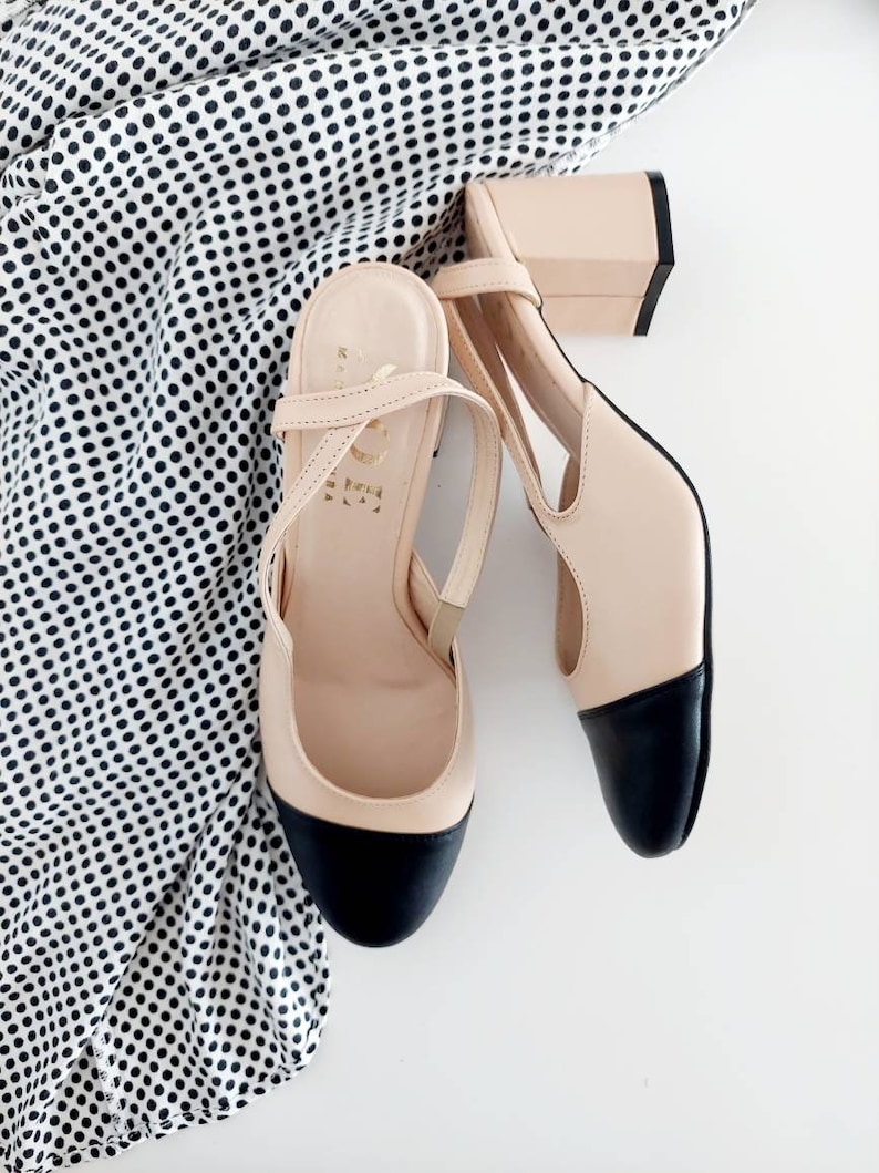 May include: A pair of beige heeled shoes with black toe caps and ankle straps. The shoes are on a white surface with a black and white polka dot fabric.