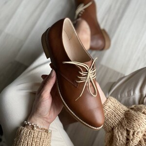 Può includere: Scarpe marroni in pelle con lacci e lacci di colore crema. Le scarpe sono tenute da una persona che indossa un maglione beige e pantaloni.