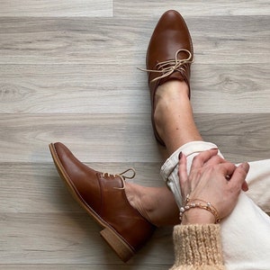 Può includere: Un paio di scarpe marroni in pelle con lacci e tacco basso. Le scarpe sono indossate da una persona con pantaloni bianchi e un maglione marrone.