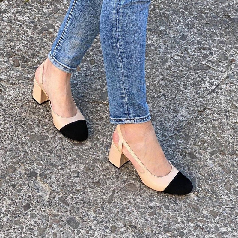 May include: A pair of beige heeled shoes with black toe caps. The shoes are slingbacks with a strap that goes around the ankle.