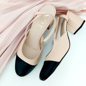 May include: A pair of beige heeled shoes with black toe caps and ankle straps. The shoes are on a white background.