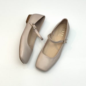 Könnte beinhalten: Ein Paar hellbeige Ballerinas aus Leder mit einem Riemen und einer Schnalle. Die Ballerinas haben eine quadratische Spitze und einen niedrigen Absatz.