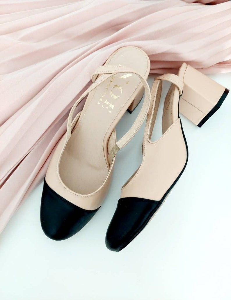 May include: A pair of beige heeled shoes with black toe caps. The shoes have a slingback strap and a block heel.