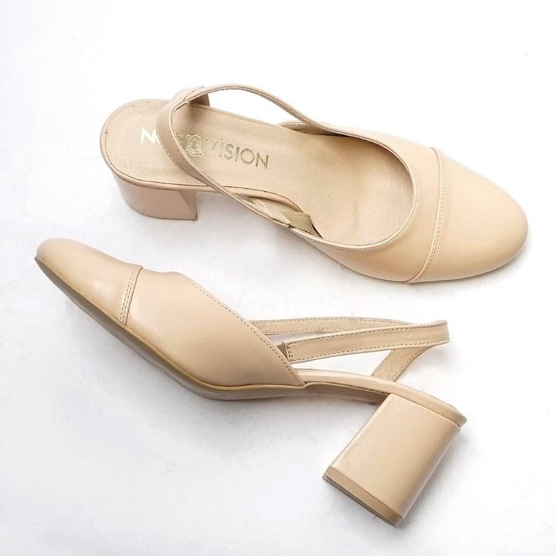 May include: A pair of beige heeled slingback shoes with a block heel. The shoes have a strap that goes around the ankle and a buckle closure. The brand name "MYVISION" is printed on the insole.