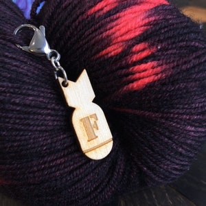 May include: A wooden bomb-shaped charm with the letter "F" engraved, connected to a silver clasp. The charm is set against a skein of yarn with a gradient of dark purple, red, and black colours.