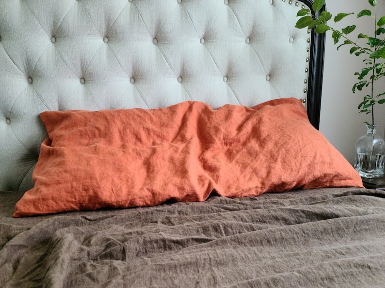 K&ouml;nnte beinhalten: Nahaufnahme eines Bettes mit einem braunen Leinenlaken und einem verbrannten orangefarbenen Kissen. Das Kopfteil ist mit einem hellbeigen Stoff mit Knopfheftung bezogen.