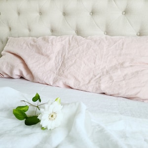 K&ouml;nnte beinhalten: Ein rosa Leinenkissen liegt auf einem wei&szlig;en Leinenbett mit einer wei&szlig;en Blume auf dem Bett.