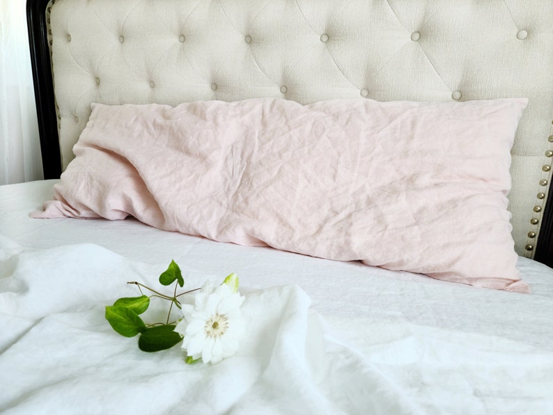 K&ouml;nnte beinhalten: Ein rosa Leinenkissen liegt auf einem wei&szlig;en Leinenbett mit einer wei&szlig;en Blume auf dem Bett.