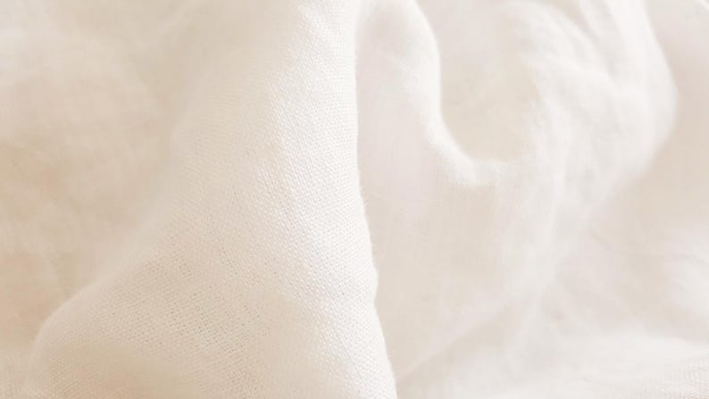 K&ouml;nnte beinhalten: Nahaufnahme eines wei&szlig;en Leinenstoffs mit einer weichen, strukturierten Oberfl&auml;che. Der Stoff ist gefaltet und drapiert und erzeugt einen glatten, flie&szlig;enden Effekt.