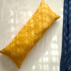 May include: A rectangular mustard yellow pillow with a textured fabric surface, positioned diagonally on a white sheet next to a folded dark blue waffle-weave blanket. The pillow has a soft, slightly wrinkled appearance with light patterns.