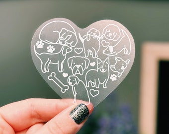 May include: A clear heart-shaped sticker with a white outline of multiple dogs and a bone. The sticker is being held by a hand with glitter nail polish.