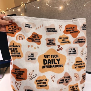 May include: A white canvas tote bag with a colorful print of affirmations for veterinary technicians. The bag features a variety of illustrations, including hearts, flowers, and rainbows. The text on the bag includes phrases such as "Vet Tech Daily Affirmations", "I Trust Myself", "I Excel at Problem Solving", and "I Make a Difference".