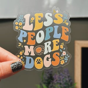 May include: A clear sticker with a colorful retro font design that reads "Less People More Dogs". The sticker is decorated with flowers and a paw print.