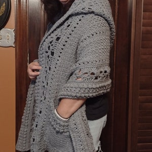 May include: A gray crocheted cardigan with a hood and a pocket. The cardigan is open in the front and has a lacy design.