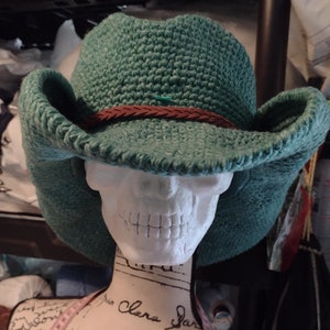 May include: A green crocheted cowboy hat with a brown leather band. The hat is sitting on a white mannequin head.