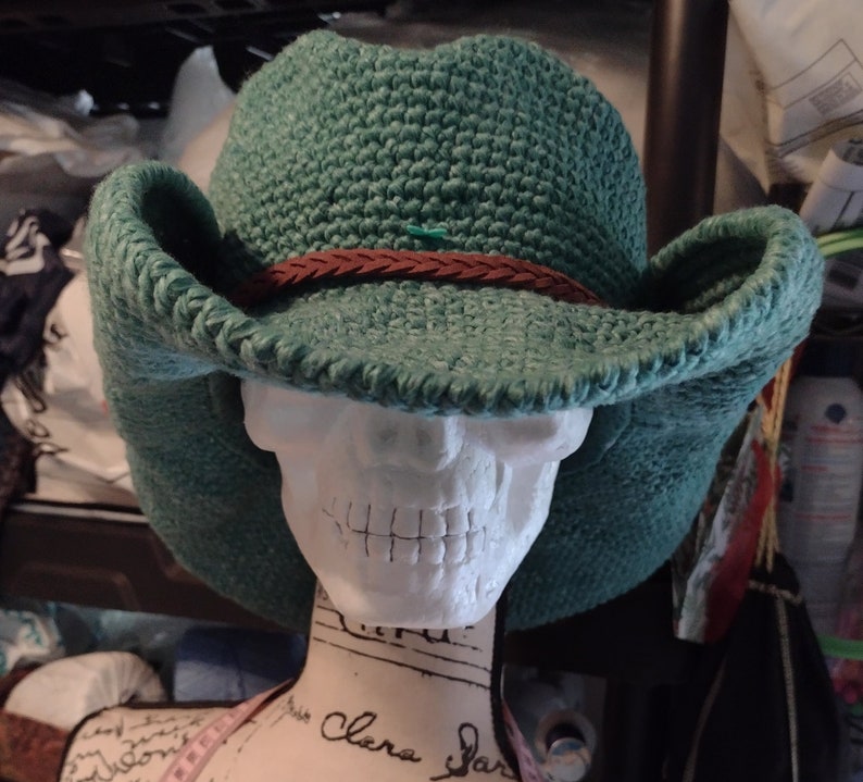 May include: A green crocheted cowboy hat with a brown leather band. The hat is sitting on a white mannequin head.