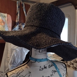 May include: A gray crocheted sun hat with a wide brim. The hat is sitting on a mannequin with a blue and white measuring tape around its neck.