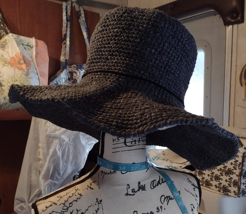 May include: A gray crocheted sun hat with a wide brim. The hat is sitting on a mannequin with a blue and white measuring tape around its neck.