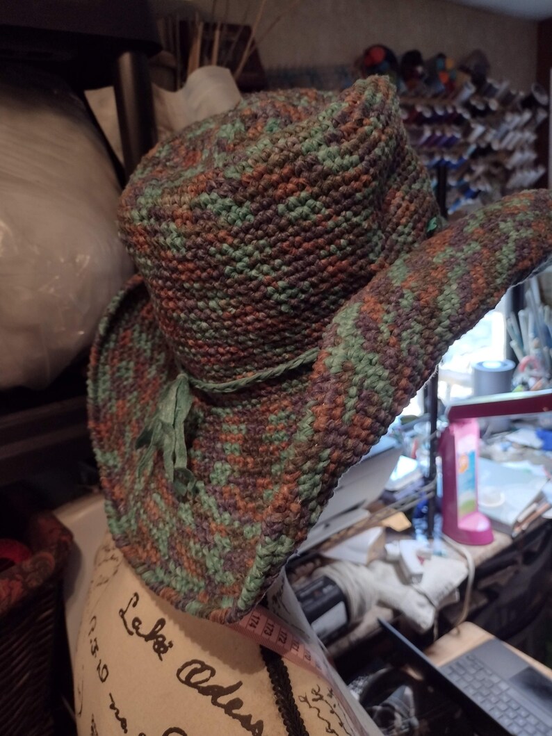 May include: A crocheted hat with a wide brim in shades of green, brown, and purple. The hat has a green ribbon tied around the crown.