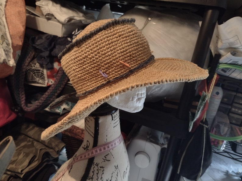 May include: A brown crocheted sun hat with a wide brim and a black trim. The hat is on a white mannequin head.