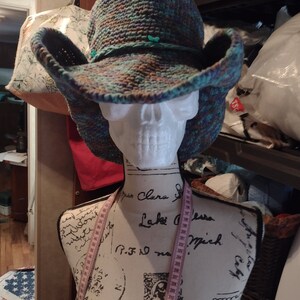 May include: A crocheted cowboy hat with a wide brim, featuring a multi-colored pattern of blue, green, and brown. The hat is displayed on a white mannequin with a measuring tape wrapped around it.