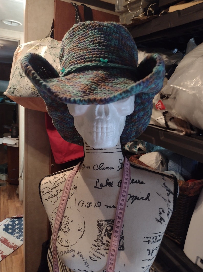 May include: A crocheted cowboy hat with a wide brim, featuring a multi-colored pattern of blue, green, and brown. The hat is displayed on a white mannequin with a measuring tape wrapped around it.