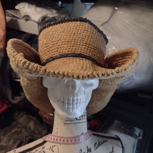 May include: A tan crocheted cowboy hat with a black band. The hat is on a white mannequin head with a pink measuring tape around the neck.