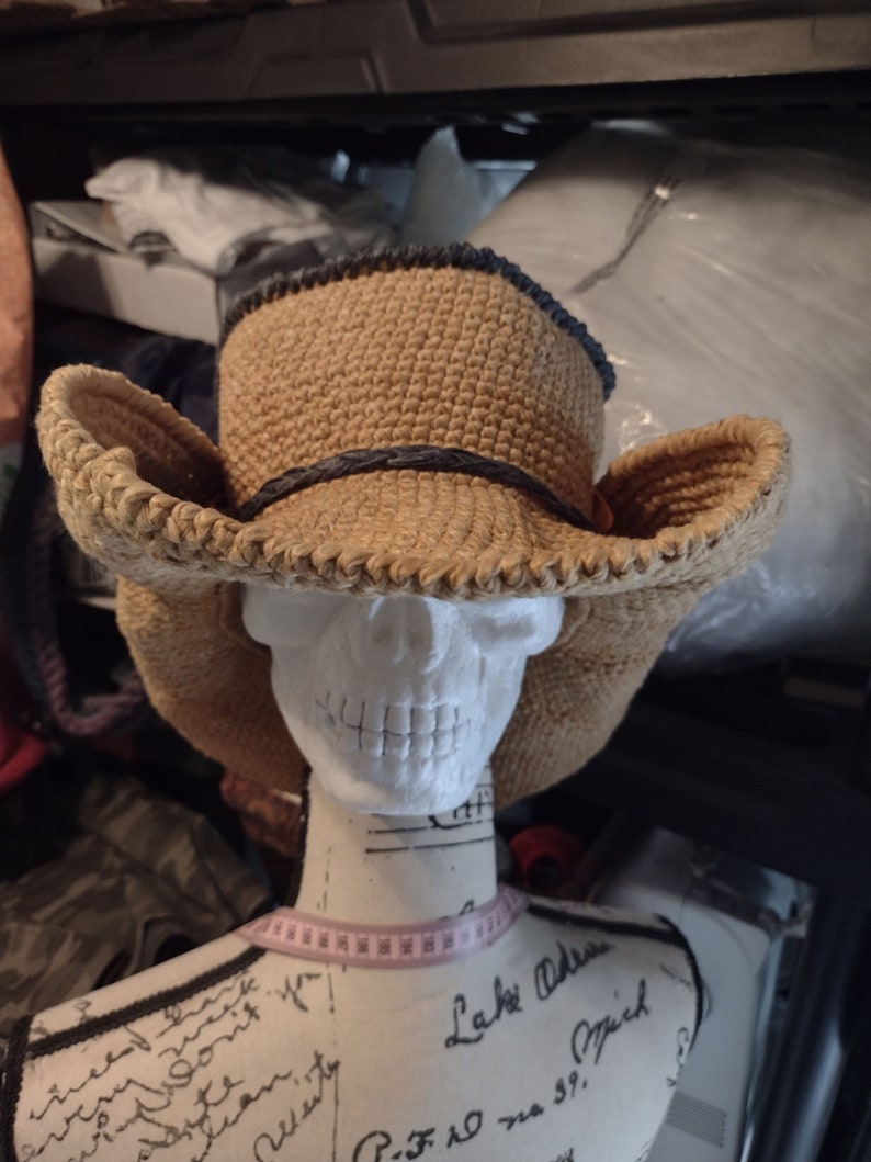 May include: A tan crocheted cowboy hat with a black band. The hat is on a white mannequin head with a pink measuring tape around the neck.
