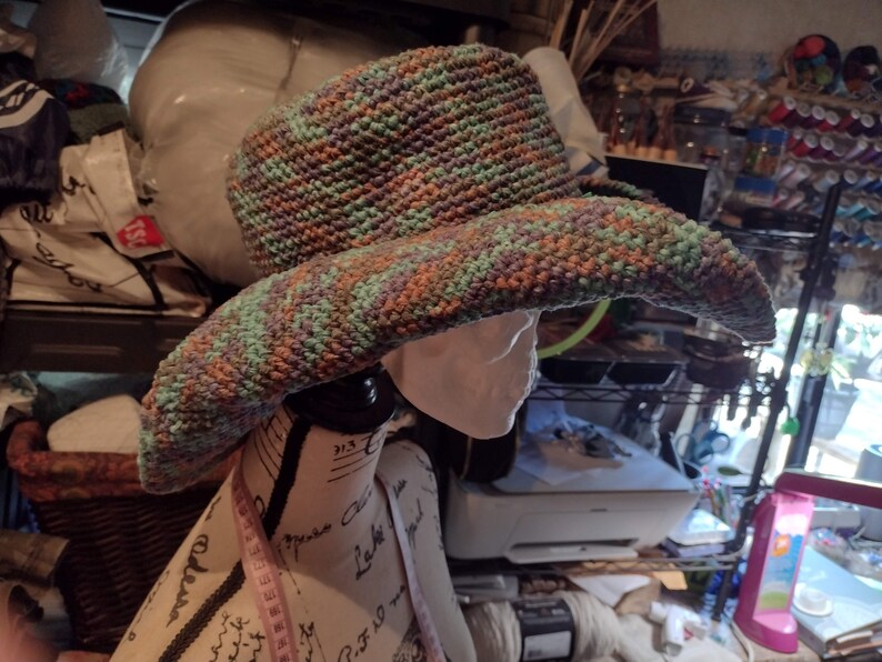 May include: A wide-brimmed crocheted hat with a green, brown, and purple color pattern. The hat is on a mannequin head.