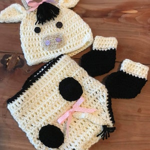 May include: A crocheted cow costume for a baby, featuring a hat with ears, a onesie with black spots, and matching mittens. The costume is made in cream and black yarn with pink accents.