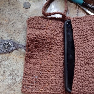 Op de afbeelding: Een bruine gehaakte telefoonhoes met een grijze gehaakte hand en een knoop. De hoes is open en een zwarte telefoon zit erin.