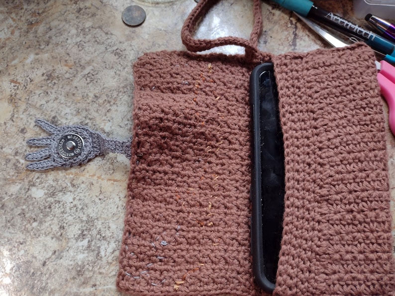 Op de afbeelding: Een bruine gehaakte telefoonhoes met een grijze gehaakte hand en een knoop. De hoes is open en een zwarte telefoon zit erin.