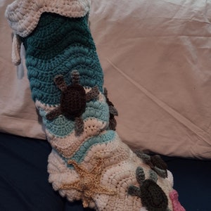 May include: A crocheted stocking with a teal, white, and brown color scheme. The stocking is decorated with crocheted sea turtles, a starfish, and white pom-poms.