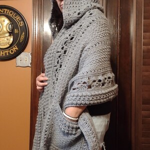May include: A gray crocheted hooded shawl with a pocket. The shawl is made with a textured stitch and has a loose, flowing design.