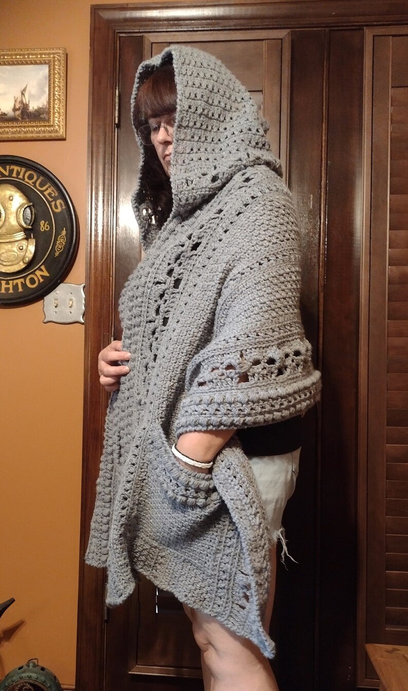 May include: A gray crocheted hooded shawl with a pocket. The shawl is made with a textured stitch and has a loose, flowing design.
