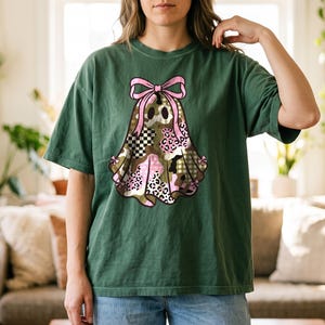 Op de afbeelding: Een donkergroen T-shirt met korte mouwen en een centrale afbeelding van een spookfiguur. Het spook is versierd met een roze strik aan de bovenkant en een patchworkpatroon van camouflage, luipaardprint en zwart-witte ruitjes.
