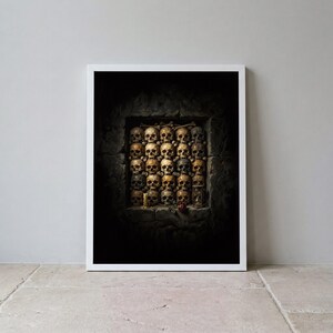 May include: A framed wall art print featuring a dark, stone-walled alcove filled with a grid of human skulls. A single red rose, a small lit candle, and a glass hourglass are positioned at the base of the skull arrangement in the dim lighting.