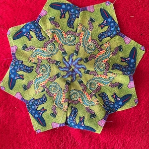 May include: A star-shaped fabric piece with a green background featuring blue and multicolored animal figures. The center has a blue floral design. The piece is set against a red background.