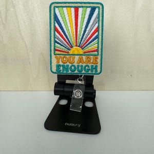 May include: A teal blue embroidered patch with a rainbow sun and the text "YOU ARE ENOUGH" in yellow and orange lettering. The patch is attached to a black retractable badge holder.