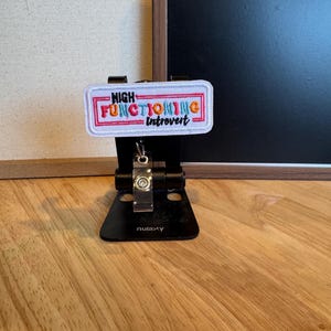 Könnte beinhalten: Ein weißes Patch mit dem Text "HIGH FUNCTIONING introvert" in bunten Buchstaben, umrahmt von einem rosa Rand. Das Patch ist auf einem schwarzen Telefonständer mit silbernem Scharnier befestigt und liegt auf einer Holzoberfläche.